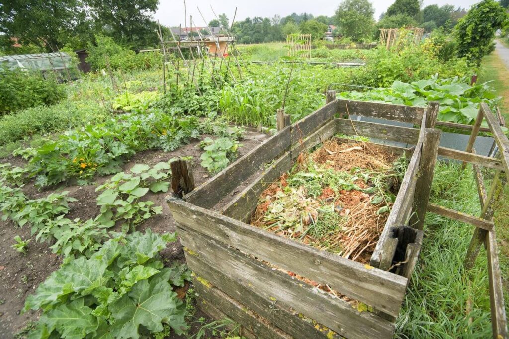 A wooden compost bin that is filled with vegetation and other yard waste and is located next to a garden